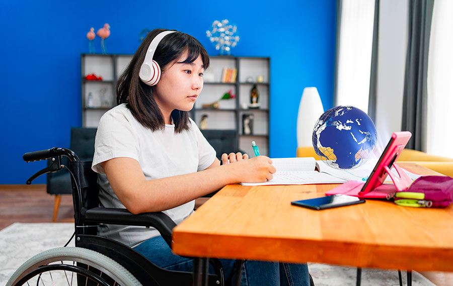 Ein Kind sitzt im Rollstuhl am Schreibtisch und notiert sich etwas auf einen Block. Es schaut auf ein Tablet und hat Kopfhörer auf.